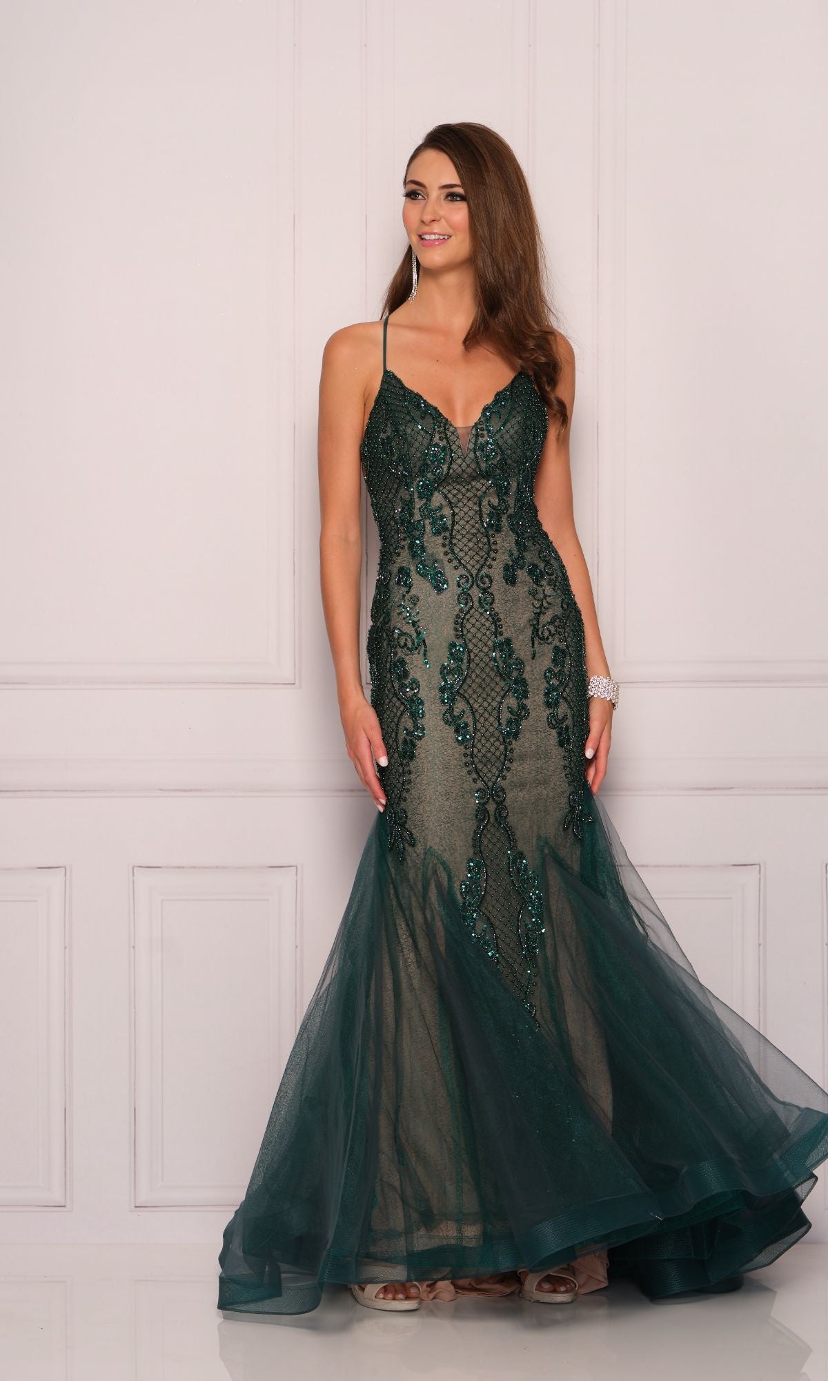 A woman models the Dave and Johnny Long Emerald Green Prom Dress 11092, a designer formal gown with lace and sequin details, standing against a white paneled wall.